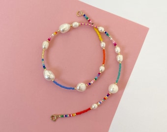 May include: A colourful beaded necklace with white pearl accents. The necklace features a rainbow of colours, including red, orange, yellow, green, blue, and purple.