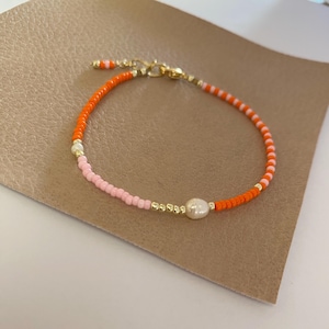 May include: A delicate bracelet made with small orange and pink beads and a single pearl accent.