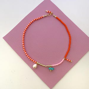 May include: A beaded necklace with orange and pink beads, a pearl charm, and a turquoise charm. The necklace is on a pink background.