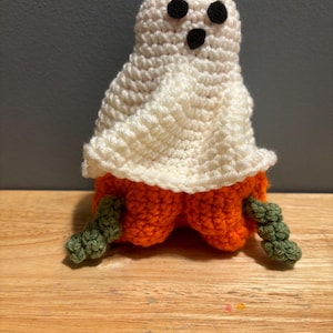 Pode incluir: Boneca fantasma de croch&ecirc; feita &agrave; m&atilde;o. O fantasma tem um corpo branco, olhos e boca pretos e uma base de ab&oacute;bora laranja. Hastes verdes est&atilde;o presas. Decora&ccedil;&atilde;o de Halloween.