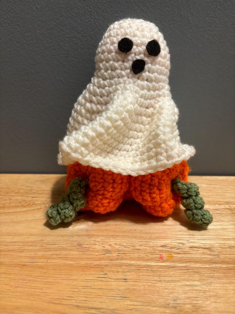Pode incluir: Boneca fantasma de croch&ecirc; feita &agrave; m&atilde;o. O fantasma tem um corpo branco, olhos e boca pretos e uma base de ab&oacute;bora laranja. Hastes verdes est&atilde;o presas. Decora&ccedil;&atilde;o de Halloween.