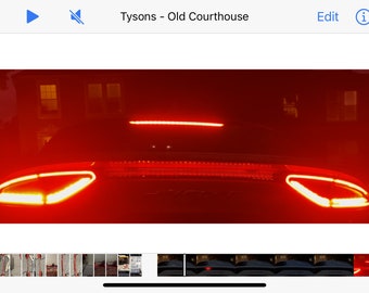 May include: A red car with its taillights on at night. The car is parked in front of a building with windows. The taillights are bright and illuminate the car's rear end.
