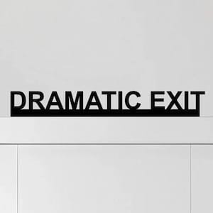 May include: A black sign with the words "DRAMATIC EXIT" above a white door. The sign is a simple, bold font, and the door is plain with a rectangular panel. The background is a plain white wall.
