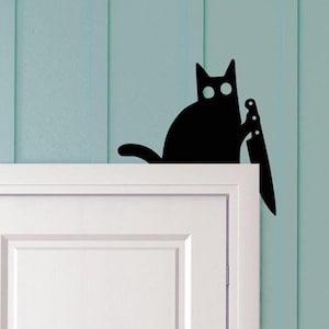 May include: Black cat silhouette holding a knife, perched above a white door frame. The cat has large, round eyes and a curved tail. The background is a light blue wall with vertical paneling.