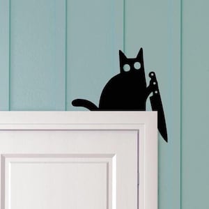 May include: A black cat silhouette holding a knife, perched atop a white door frame. The cat has large, round eyes and a curved tail. The background is a light blue wall with vertical paneling.