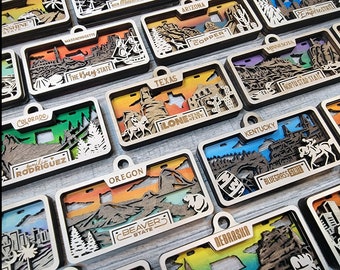 May include: A collection of laser-cut wooden state-themed ornaments. Each ornament features a layered design with state names and iconic imagery, such as mountains, cacti, and city skylines. The ornaments have a natural wood finish with colorful backgrounds.