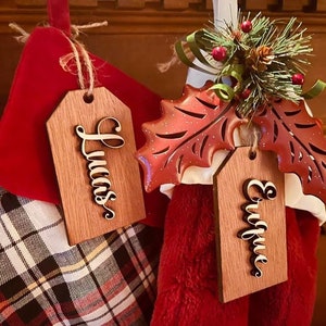 May include: Two Christmas stockings with wooden name tags. One tag says "Lucas" and the other says "Enrique". The stockings are red and white, with a plaid pattern on one. A decorative holly leaf with berries and pine needles is also visible.