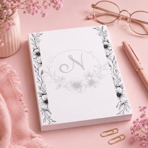 Op de afbeelding: Een witte notitieblok met een bloemenrand en een grijze letter "N" in een bloemenkrans. Het notitieblok staat tegen een roze achtergrond, met een roze pen, gouden paperclips en een bril.