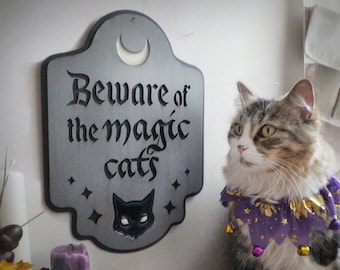 May include: A silver metal sign with the text "Beware of the magic cats" in black lettering. A crescent moon is in the upper left corner of the sign. A black cat silhouette is in the lower center of the sign. A cat wearing a purple and gold jester costume is sitting in front of the sign.