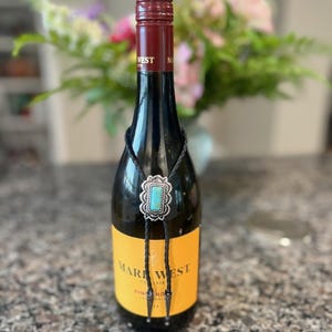 May include: A bolo tie featuring a rectangular turquoise stone set in a scalloped silver-tone metal frame with a braided black leather cord and silver-tone metal tips, displayed around the neck of a Mark West Pinot Noir wine bottle.