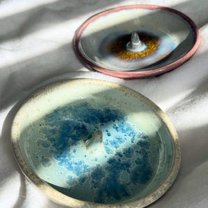 Peut inclure: Deux coupelles à bagues rondes en céramique avec des cônes centraux. La coupelle avant présente une base vert clair avec des motifs de glaçure cristalline bleue. Celle de derrière a un centre blanc, un anneau brun foncé et un bord rose.