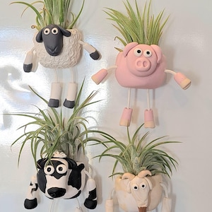 May include: Four whimsical animal-shaped planters, each holding a green air plant. The planters are a sheep, pig, cow, and goat, with dangling legs and arms. The sheep is white with a black face, the pig is pink, the cow is black and white, and the goat is beige.