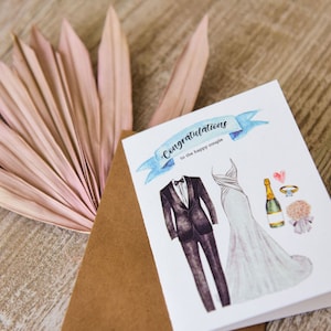 May include: A wedding congratulations card with a watercolour illustration. The card shows a tuxedo, a wedding dress, a champagne bottle, a ring, and a bouquet. The text on the card reads "Congratulations to the happy couple."