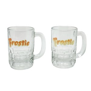May include: Two clear glass mugs with handles. The word "Frostie" is printed in orange letters on the front of each mug. The mugs have a textured, paneled design on the lower half and are suitable for beverages.