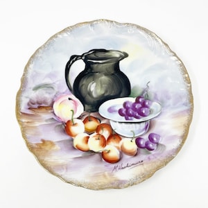 May include: A decorative, hand-painted plate featuring a still life of fruit and a pitcher. The plate has a scalloped edge with gold trim. The artwork depicts a black pitcher, peaches, apples, and grapes. The artist's signature, "M. Hashimura," is visible.