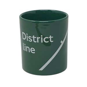 Puede incluir: Una taza de cerámica de color verde oscuro con el texto blanco District line en la parte frontal, acompañada de un gráfico de línea diagonal blanca que se extiende por el lateral del recipiente cilíndrico, creando un diseño limpio y sencillo.