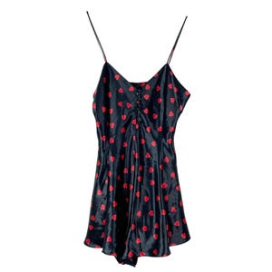 Könnte beinhalten: Schwarzer Satin-Romper mit rotem Herzmuster. Er hat Spaghettiträger, einen V-Ausschnitt und eine Knopfleiste vorne. Das Kleidungsstück hat einen ausgestellten Rock und ist für eine bequeme Passform konzipiert. Der Stoff hat einen leichten Glanz.
