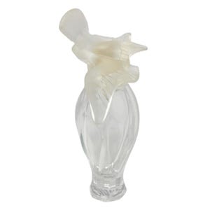May include: A clear glass perfume bottle with a frosted glass bird design on top. The bottle has a fluted, teardrop shape. The bird is depicted in flight, with outstretched wings. The bottle is empty and sits against a white background.
