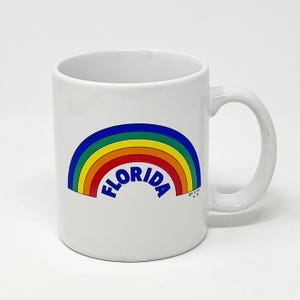 Könnte beinhalten: Weißer Keramikbecher mit einem bunten Regenbogen-Design und dem Wort "FLORIDA" in Blau. Der Regenbogen hat rote, orange, gelbe, grüne und blaue Bänder. Der Becher hat einen gebogenen Griff und ist für heiße oder kalte Getränke geeignet.