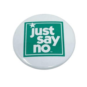 May include: A circular white pinback button featuring a green square border. Inside the square, the text "just say no" is printed in white, lowercase letters, accompanied by a small white star in the top left corner and a small TM symbol.