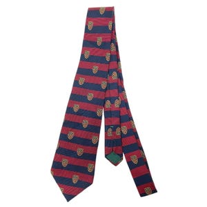 May include: A necktie featuring horizontal stripes in navy blue and red. The fabric is decorated with a repeating pattern of small, multi-colored crests or shields. The tie has a pointed end and a visible green fabric label on the inner lining.