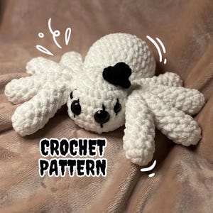 Peut inclure: Araignée blanche en crochet avec des yeux noirs et un nœud papillon noir. L'araignée est posée sur une couverture rose. Le texte "CROCHET PATTERN" est écrit sous l'araignée.