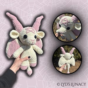 May include: A white crocheted goat with pink wings and horns. The goat has a pentagram on its forehead and large black eyes. The goat is being held by a hand.