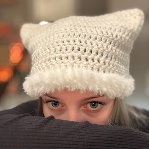 May include: A cream-colored crochet hat with pointed ears and a fluffy white trim. The hat is made of a textured yarn and is designed to be worn on the head. The hat is a unique and stylish accessory.