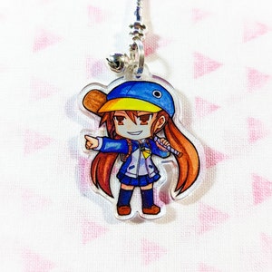 Puede incluir: Un charm de acrílico transparente con una niña de dibujos animados de pelo largo y castaño, que lleva una gorra de béisbol azul y amarilla, una chaqueta azul y una falda azul. Ella sostiene un micrófono y apunta hacia la derecha.