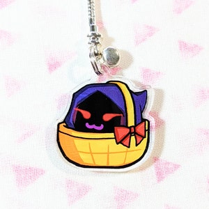 May include: A purple and black character with red eyes and a pink bow is inside a yellow basket. The character is wearing a purple hood. The basket is hanging from a silver chain.