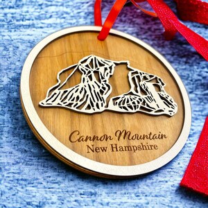Könnte beinhalten: Ein Holz-Anhänger in Form des Cannon Mountain in New Hampshire. Der Anhänger ist braun und hat einen weißen Rand. Er ist mit einem roten Band verziert.