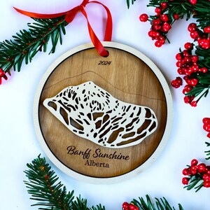 May include: A wooden Christmas ornament featuring a laser-cut design of a mountain range with the text "Banff Sunshine Alberta" and the year "2024".