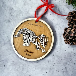 May include: A round wooden Christmas ornament with a map of Okemo, Vermont, cut out in white. The ornament has a red ribbon for hanging and the text "WOOD FAMILY 2023".