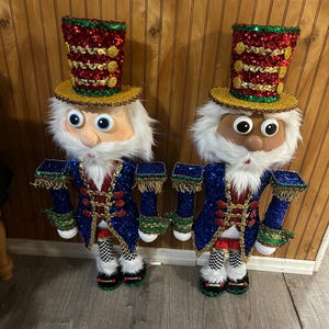 Op de afbeelding: Twee decoratieve notenkrakerfiguren met blauwe jasjes vol lovertjes, rood-gouden hoeden en wit imitatiebont als haar en baard. Elke figuur draagt een zwart-wit geblokte legging, gouden franje-epauletten en zwarte schoenen met een groene rand.