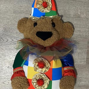 Op de afbeelding: Een bruine teddybeer verkleed als een clown. De beer draagt een kleurrijke patchwork-outfit met rode en gouden accenten, een gerimpelde kraag en een puntmuts. De zwarte schoenen van de beer hebben gouden veters.