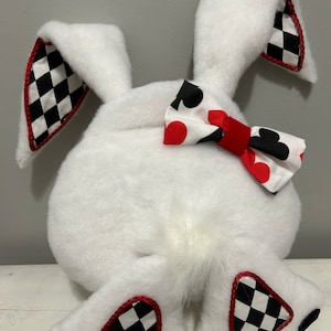 以下が含まれることがあります： 白のぬいぐるみウサギ。蝶ネクタイと、白黒のチェック柄と赤いトリムがアクセントになっています。蝶ネクタイには赤いハートと黒のスペードの模様があります。ウサギの耳と足にもチェック柄のアクセントがあります。