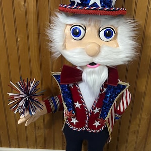 May include: A patriotic Uncle Sam doll with a red, white, and blue color scheme. The doll wears a top hat, a bow tie, and a star-studded jacket. It holds a decorative starburst in its hand. The doll's shoes are black and white.