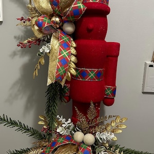 May include: A red velvet Christmas nutcracker figurine with a gold and blue plaid ribbon and gold glitter accents. The nutcracker is surrounded by greenery, gold glitter sprigs, and white berries.