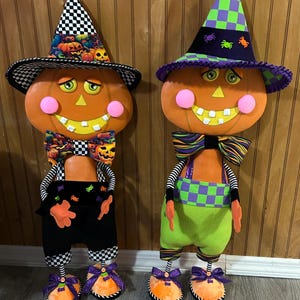 May include: Two decorative pumpkin figures with smiling faces, pink cheeks, and square teeth. The left figure wears a black and white chequered hat and bow tie with pumpkin print, while the right figure wears a purple and green chequered hat and bow tie.