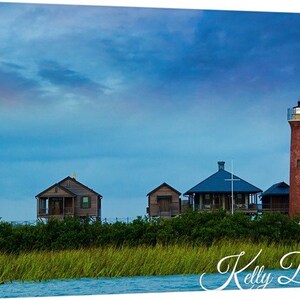 K&ouml;nnte beinhalten: Ein roter Backstein-Leuchtturm ragt hoch in einen blauen Himmel mit ein paar Wolken.  Im Vordergrund befinden sich mehrere Holzh&auml;user, im unteren Bildbereich gr&uuml;nes Gras und Wasser.  Der Text "Kelly De Long" ist unten im Bild in Kursivschrift geschrieben.