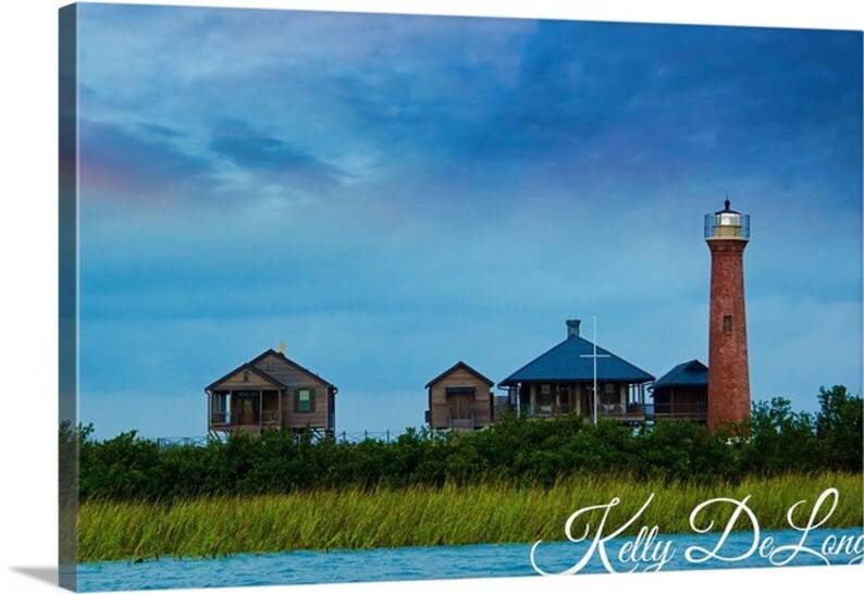 K&ouml;nnte beinhalten: Ein roter Backstein-Leuchtturm ragt hoch in einen blauen Himmel mit ein paar Wolken.  Im Vordergrund befinden sich mehrere Holzh&auml;user, im unteren Bildbereich gr&uuml;nes Gras und Wasser.  Der Text "Kelly De Long" ist unten im Bild in Kursivschrift geschrieben.