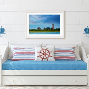 K&ouml;nnte beinhalten: Ein wei&szlig;es Daybed mit einer blauen Matratze und rot-wei&szlig; gestreiften Kissen. &Uuml;ber dem Bett h&auml;ngt ein gerahmtes Foto eines Leuchtturms und eines bew&ouml;lkten Himmels. Zwei blaue Wandleuchten mit k&auml;figartigen Schirmen befinden sich auf beiden Seiten des Bildes.