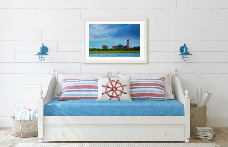 K&ouml;nnte beinhalten: Ein wei&szlig;es Daybed mit einer blauen Matratze und rot-wei&szlig; gestreiften Kissen. &Uuml;ber dem Bett h&auml;ngt ein gerahmtes Foto eines Leuchtturms und eines bew&ouml;lkten Himmels. Zwei blaue Wandleuchten mit k&auml;figartigen Schirmen befinden sich auf beiden Seiten des Bildes.