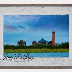 K&ouml;nnte beinhalten: Ein gerahmtes Foto eines roten Leuchtturms auf einer kleinen Insel mit blauem Himmel und Wolken. Im Vordergrund befinden sich drei kleine H&auml;user und im Vordergrund ein Gew&auml;sser. Der Name des Fotografen, Kelly De Long, ist unten im Bild in Kursivschrift geschrieben.