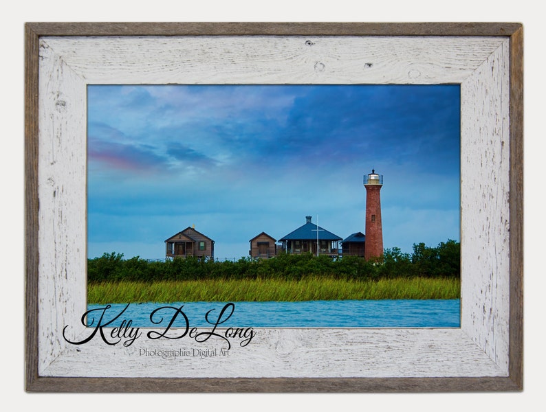 K&ouml;nnte beinhalten: Ein gerahmtes Foto eines roten Leuchtturms auf einer kleinen Insel mit blauem Himmel und Wolken. Im Vordergrund befinden sich drei kleine H&auml;user und im Vordergrund ein Gew&auml;sser. Der Name des Fotografen, Kelly De Long, ist unten im Bild in Kursivschrift geschrieben.