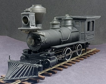 May include: A detailed model of a black steam locomotive with white-rimmed wheels, resting on brown railroad tracks. The model features a headlight, smokestack, and a small cabin. The locomotive is a miniature replica of a classic train.