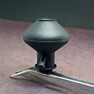 May include: A black 3D printed model of a water tower with a round top and a cylindrical base. The model is being held by a pair of tweezers.