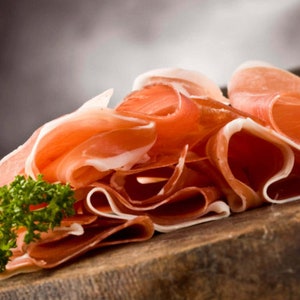 May include: Thinly sliced prosciutto arranged on a wooden surface, with a sprig of fresh parsley. The cured meat has a reddish-pink color with white marbling, and is slightly curled.