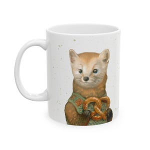 May include: White ceramic mug featuring a watercolor illustration of a cute pine marten wearing a green sweater and holding a pretzel. The marten has brown fur and blue eyes. The mug has a white handle and is decorated with green paint splatters.