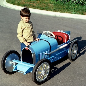 May include: A young boy stands next to a blue toy car that resembles a classic Bugatti. The car has a red leather seat and a steering wheel. The boy is wearing a tan jacket and blue jeans.